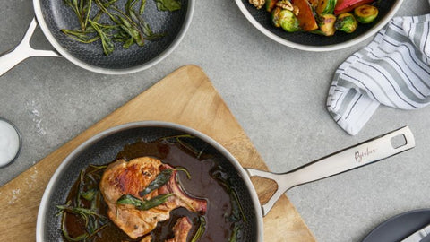Pork chops with herbs in a pan on a wooden board, surrounded by ingredients and cutlery.